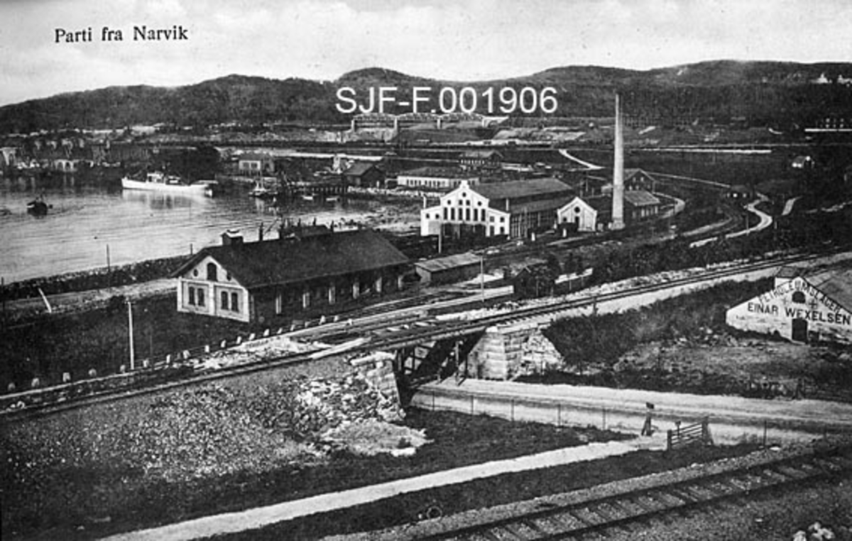 Fra jernbane- og kaiområdet i Narvik i Nordland.  I forgrunnen krysser en veg to jernbanespor gjennom underganger.  Mellom og nedenfor jernbanetraseene står det murbygninger, antakelig reist med tanke på industrielle funksjoner.  Jernbanelinjene og en nedenforliggende vegtrase følger i grove trekk den buete linja til bukta til venstre i bildet.  Her ligger et par skip, antakelig i påvente av malmlasting. 