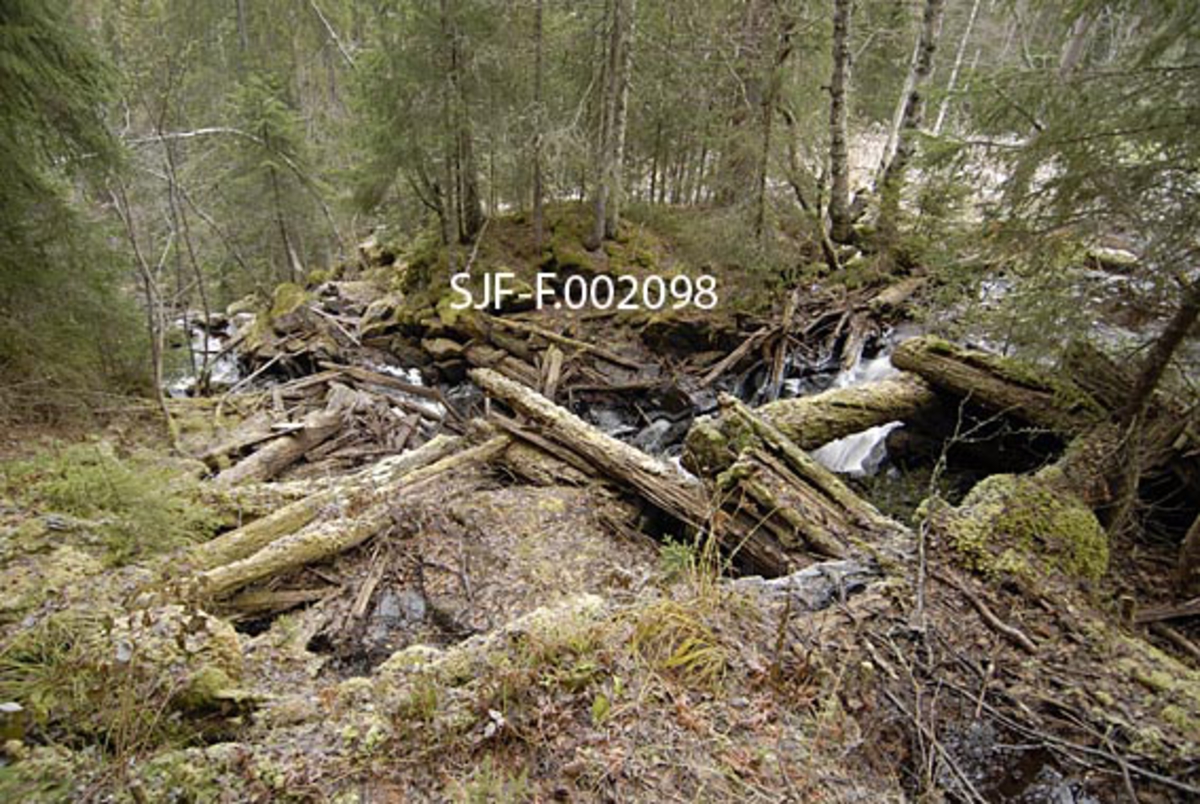 Fra Sagfallet i det lille vassdraget Anda (uttales Ana) på Storsjøens østside i Ytre Rendalen.  Her renner elva bratt nedover gjennom et berglandskap før den 70-80 meter lavere når den nedenforliggende innsjøen like sør for bruket Andråstøa.  Øverst i dette fallet lå Andrå-gardenes oppgangssag.  Høsten 1963 ble denne saga, i sterkt forfallen tilstand, demontert og flyttet til det daværende Norsk Skogmuseum i Elverum. Fotografiet er tatt øverst i Sagfallet, omtrent der den øvre delen av saghuset må ha stått.  Vassrenna ned til underfallshjulet som drev anlegget må ha gått bratt nedover fra et tømmerfundament bak den lille grana helt til høyre i bildet.  En del råtne tømmerstokker som ligger i forgrunnen var antakelig komponenter fra saghuset som allerede tidlig i 1960-åra var så ødelagte at de ikke kunne brukes ved gjenoppføring av anlegget på museet.  Dette virket ble av den grunn forlatt på sagtomta.  På en liten haug på motsatt side av fallet, om lag midt i fotografiet, skimtes en liten inngravning i terrenget, hvor det later til å ha stått et kvernhus.