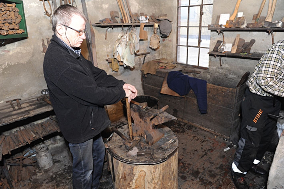 John Lystad (f. 1945) fra Drammen, fotografert i Knudsen-smia i Mjøndalen, der han som hobby arbeidet som oppslager og medhjelper for øksesmeden Oddbjørn Knudsen, som vi skimter helt til høyre i dette fotografiet.  Lystad står ved stabben med ambolten og støtter seg på skaftet til en tung hammer han er klar til å bruke under ”høling” av merkedelen til ei merkeøks.  At han venter på denne måten skyldes at Knudsen måtte gi arbeidsstykket ei oppvarming før karene kunne starte arbeidet med skaftmeisel (Knudsen) og hammer (Lystad) – jfr. SJF-F. 003354-SJF-F. 003355. 