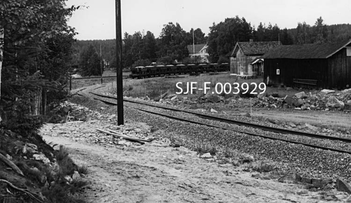 Første jernbanetransport på sidesporet mellom Geithus Bruk på Modum i Buskerud og Geithus stasjon på Randsfjordbanen.  Fotografiet er tatt mot en kurve med en bredsporet, enkel skinnegang.  Et stykke framme i kurven ser vi ei rekke med vogner (ni stykker) som antakelig var lastet med pukkstein som skuller brukes på anleggsområdet for den nye bedriften.  På motsatt side av jernbanetraséen, til høyre i bildet, lå et litt malingsslitt småbruk.  Lengre nede, bak vognrekka, skimter vi en noe større, kvitmalt sveitserstilvilla omgitt av en hage med store lauvtrær. 