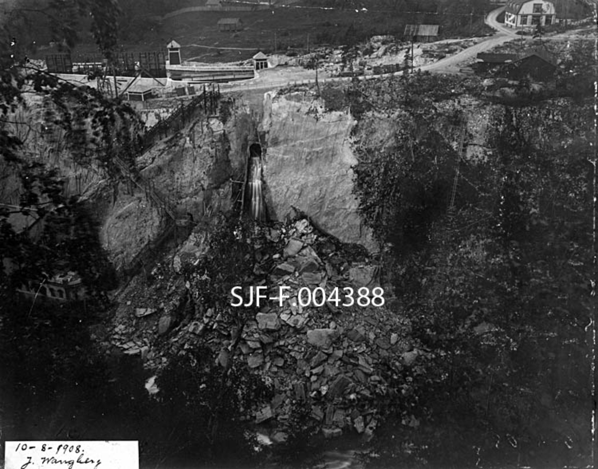 Fra elvegjelet som omgav den nedre delen av Tinnåa i Notodden (da fotografiet ble tatt i «Hiterdal» eller Heddal) kommune i Telemark, slik det så ut 10. august 1908. Fotografiet er tatt fra platået på østsida av elvegjelet, mot industriområdet til Tinfos Papirfabrik på vestsida.  Sentralt i bildet ser vi en ”tipp” – en tunnelåpning i berget med et nedenforliggende skråplan av tømmer, hvor det later til å være tippet ned en masse sprengstein, dels svært store blokker, som er blitt liggende ved foten av det bratte gjeldet, mot elveleiet, som vi her bare så vidt skimter nederst i bildet.  Til venstre i bildeflata ser vi trappa fra vegen og industriområdet oppe på platået ned mot kraftstasjonen som Tinfos Papirfabrik satte i drift nede ved elvas vestre bredd i 1901.  Kapasiteten ved kraftstasjonen, og dermed vannforbruket, ble utvidet i 1904 og 1907.  Etter den sistnevnte utbygginga oppsto det en stømvirvel – ”Tinnfoss-sveiva” – i vassdraget, som ifølge Skiensvassdragets Fellesfløtningsforening bidro til brudd- og sprekkskader, samt slitasje, på tømmeret, noe som reduserte anvendelsesmulighetene og dermed verdien til virket.  Saken ble brakt inn for retten.  Advokaten til Tinfos Papirfabrik la fram dette bildet og flere andre (SJF-F. 004384 - SJF-F. 004416) fra samme del av vassdragets som bevismateriale i en skjønnssaken. Saken ble ført i Nedre Telemark sorenskriveri i perioden 1907-11, deretter som ankesak i Bergen overrett fram til 1914 og til slutt i Høyesteretts kommisjon for skriftlige saker i 1916-17.  Poenget med fotografiene skal dels ha vært å vise at Tinfos Papirfabrikk ikke var aleine om å ha tipppet steinmasser fra anleggsarbeid i Tinnfossen, dels å vise at flomvannet førte mye slik masse med seg videre nedover, og dermed «renset» elveløpet slik at de negative konsekvensene for tømmerfløtinga ikke ble varige.  Dette fotografiet skulle antakelig sammenliknes med SJF-F. 004389, som ble tatt fra samme opptakssted i begynnelsen av mai samme år. 