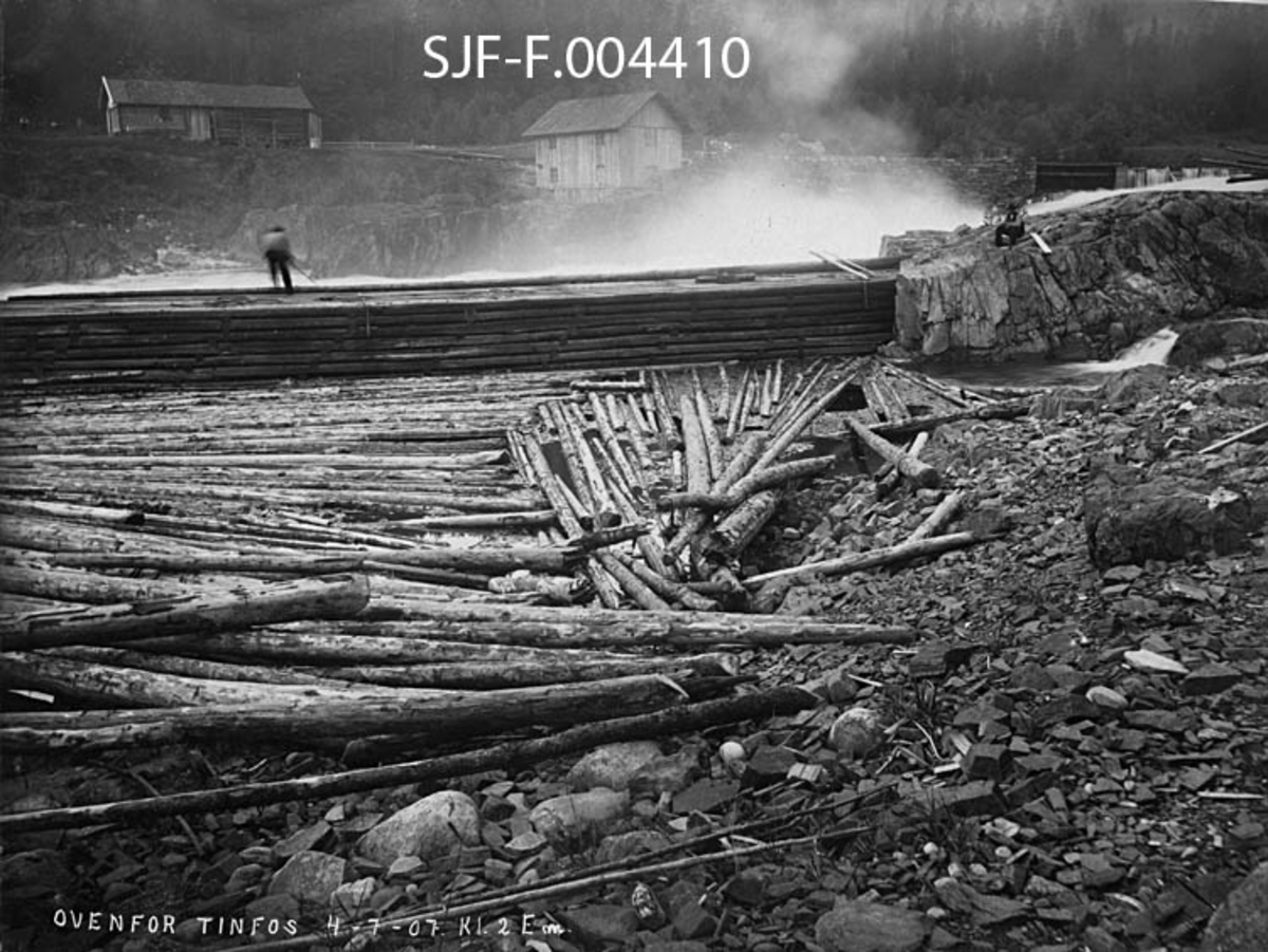 Fløtingsaktivitet ved Sagafoss i Tinnåa i Notodden kommune i Telemark (da dette fotografiet ble tatt het kommunen «Hiterdal» [Heddal]).  Bildet ble tatt 4. juli 1907 klokka 14. 00.  Fotografiet er tatt fra ei steinete strand der vi, i forgrunnen, ser en del fløtingsvirke som er blitt liggende i ei bakevje bak en skådam i tømmerkistekonstruksjon, som antakelig skulle hindre at tømmeret la mot land nettopp her.  Da bildet ble tatt sto en fløter på skådammen, med ryggen vendt mot fotografiet og haken mot den kvitskummende Sagafossen, antakelig for å hindre at virket satte seg fast.  På den bergnabben skådammen var en forlengelse av satt en kollega, kledd i svarte bukser, mørk vest, lys skjorte og med hatt på hodet på ei trekasse og kvilte litt.  Bak ham, til høyre i bildet, skimtes noe av damkonstruksjonen ved innløpet til Sagafoss.  Sentralt i den øvre delen av bildet lå en toetasjes bordkledd bygning – kanskje et mølleanlegg – like nedenfor dammen.  På elvebakken litt lengre til venstre ser vi uthusbygningen på et småbruk.  Dette fotografiet er blant de 30 opptakene Tinfos Papirfabrik leverte som bevismateriale i forbindelse med en tvist med Skiensvassdragets Fellesfløtningsforening i 1907.  Striden hadde sitt opphav i «Tinnfoss-sveiva», en strømvirvel som hadde oppstått etter at papirfabrikken foregående vinter hadde bygd ny dam, økt kapasiteten på sitt vanninntak og tippet stein fra anleggsarbeidet i vassdraget under Tinnfossen.  For advokaten til papirfabrikken var det et poeng å vise, at skader på fløtingsvirket kunne ha oppstått andre steder i vassdraget, som her.  De knekte tømmerstokkene i forgrunnen, på et fotografi som var tatt ovenfor «Tinnfoss-sveiva» ble antakelig brukt i denne argumentasjonen.  Mer om konflikten mellom Skiensvassdragets Fellesfløtningsforening og Tinfos Papirfabrik i rubrikken «Andre opplysninger». 