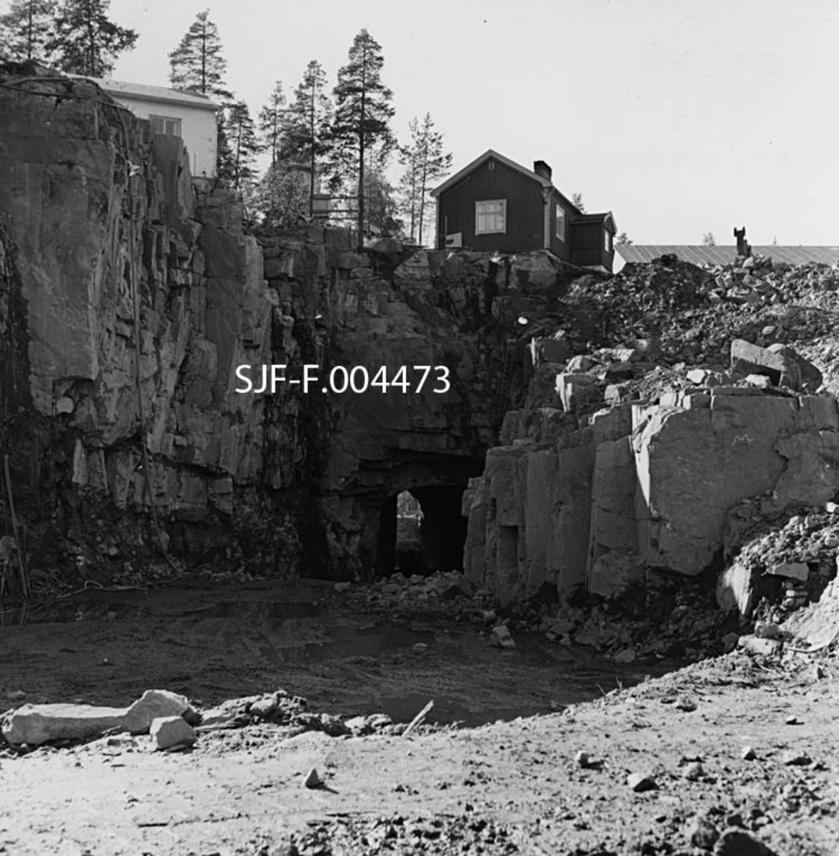 Driftstunnelen for Geithusfoss kraftverk på Modum i Buskerud, fotografert 1. September 1960, et par uker etter at anleggsmannskapene oppnådde gjennomslag.  Fotografiet er da også tatt fra en slik vinkel at vi ser lys fra den andre sida gjennom den bortimot 110 meter lange passasjen som ble sprengt i berget under industritomta til Drammenselvens Papirfabrikker.  Oppå berget ser vi også to av fabrikkens bygninger.  Det kvitmalte huset vi ser hjørnet av til venstre inneholdt rom for portvakta samt et vaskeri, mens det mørkere (antakelig rødmalte) huset over traseen for driftstunnelen ble brukt som spisebrakke.  Geithusfoss kraftverk ble satt i drift 1. oktober 1961, men arbeidet med å legge forholdene til rette for energiproduksjonen fortsatte også påfølgende vinter, da det ble reist en dam som hevet vannspeilet ovenfor fossen noe. 