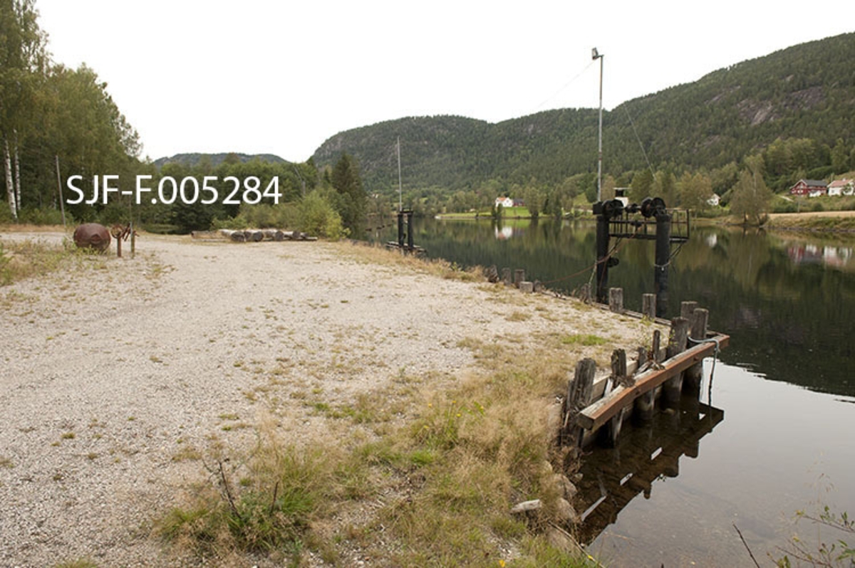 Fra tømmeropptaksplassen ved Kåsa, på vestsida av vassdraget mellom Strengen og Hogga, øverst i Bandak-Norsjø-kanalen (Nome kommune i Telemark).  Her trengte Skiensvassdragets Fellesfløtningsforening en opptaksplass for virke som hadde vært slept i «klubber» (bunter) over Vestvannene (Bandak, Kviteseidvatn og Flåvatn i Vest-Telemark) for å kjøre det med lastebil til Norsjø eller eventuelt helt fram til industrien i Skien, i første omgang mens det ble utført vedlikeholdsarbeid på de nedenforliggende sluseanleggene og bygd ny kraftverksdam ved Hogga.  Et av problemene var å finne et sted langs den smale bilvegen oppover langs kanalen der det var plass nok til å lesse opp tømmer på lastebiler uten at arbeidet var til sjenanse eller et faremoment for annen trafikk.  Dette ble gjort ved å slå ned en pålestøttet skjerm mot vassdraget og fylle opp med drenerende masser bak, mot vegen, noe vi ser på dette fotografiet.  Mye av den øvrige biltrafikken ble flyttet til en trasé som gikk over Storemoen lengre vest.  Opptaksplassen ble ferdigstilt i 1979 og hadde da kostet 415 970 kroner.  Ute i vatnet ble det reist to par krantårn i stål med en horisontal bom med to kjettingtromler på toppen.  Kjettinger fra disse tromlene ble hektet til pålene ved bryggekanten og senket slik at de lå undern vannspeilet mellom tårnene og brygga.  Deretter kunne man lede en tømmerbunt (et lastebillass) inn i dette vannbassenget, for så å stramme opp kjettingene slik at tømmeret lå støtt på dette underlaget.  Deretter kunne vaierbindene rundt buntene løsnes, og virket kunne hentes inn på lastebilene ved hjelp av kranene på disse kjøretøyene.  En kraftig stålbjelke med et par tunge lodd sørget for at kjettingene mellom tårnene i elva og bryggekanten hele tida var stramme, slik at tømmeret lå stabilt på dette underlaget inntil det var berget inn på land. 