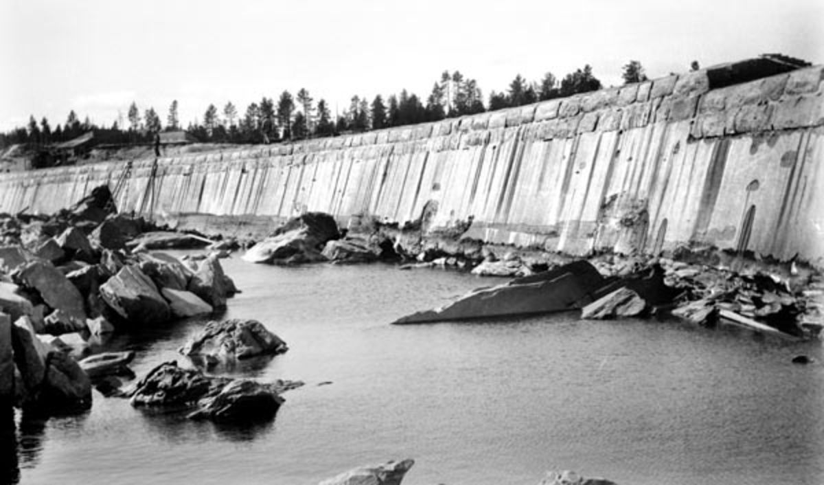 Damanlegget ved Skjefstadfossen i Glomma, sett nedenfra.  Skjefstadfossen ligger i Heradsbygda i Elverum kommune i Hedmark.  Her ble det første store kraftutbyggingsprosjektet i denne delen av Glomma-vassdraget realisert i 1909-1910.  Dette bildet er antakelig tatt like etter at dammen var ferdigstøpt, mens det ennå var meget lav vannføring i vassdraget.  Fotografen har stått nedenfor det kraftige, østre landkaret, og med kameraet vendt mot den buete fronten på dammen.  Fotografiet viser hvordan betongkonstruksjonen hadde vært forskalet.  Damkrona trappes noe ned mot øst, der man ønsket at mesteparten av fløtingstømmeret skulle passere.  Vi ser også hvordan ytterkanten på damkrona er forsterket med to floer kvadret naturstein.  Til venstre i bildet, den den delen av elvebrinken som rager over damkrona, skimter vi et par provisoriske skur som var brukt i anleggsperioden. 

Litt mer informasjon om kraftanlegget ved Skjefstadfossen finnes under fanen «Andre opplysninger». 