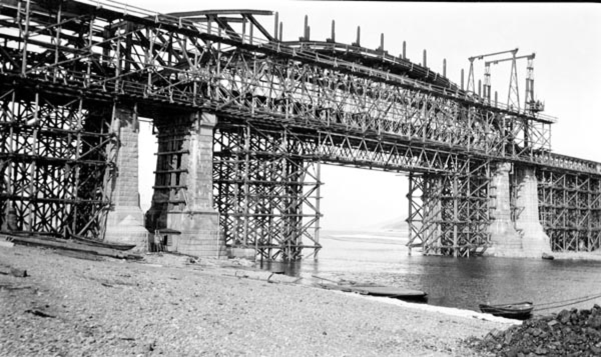 Brua over Mjøsas utløp i Vorma ved Minne, fotografert i forbindelse med ombyggingsarbeid, tidlig på 1900-tallet.  Fotografiet viser brua med stillasene som ble brukt under ombygginga.  Sjølve brua er en sammenboltet støpejernskonstruksjon (fagverksbru) som kviler på kraftige brukar av hoggen naturstein, plassert parvis på hver side av djupålen i vassdraget.  Ved stranda er det fortøyd en robåt og en bred pram.  Også inne på land, like innunder det nevnte trestillaset, ligger det en robåt. Denne brua ble avløsy av ei ny, nesten 840 meter lang jernbanebru over Minnevika, utført i betong, høsten 2023.