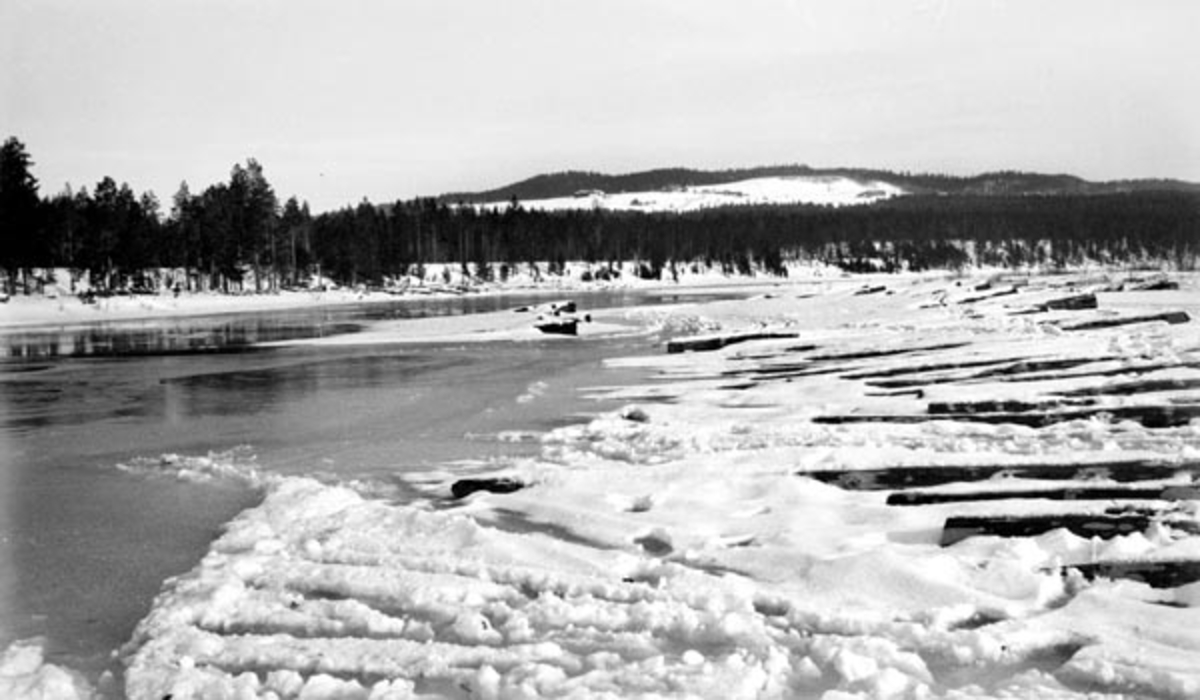 Seinvintersbilde fra Storkvernøra i den delen av Glomma som renner gjennom Tolga kommune.  Fotografiet er tatt fra en elvebredd der det til høyre ligger en del fløtingstømmer i snøen.  Til venstre er det overvann på isen.  Langs elvebreddene er det snødekte grusrygger hvor det vokser furuskog.  I bakgrunnen en åskam hvor det ligger et par gardsbruk, omgitt av snødekte jorder. 