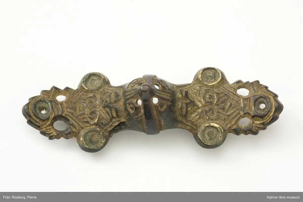 KLM 6. Spänne, likarmat spänne. Gjutet av brons. Mittornamentet gjutet i ett stycke med spännet. Vardera ändstycket har tre utstående flikar med rundlar, alla genomborrade. Spår av förgyllning. På undersidan finns nålfäste och nålhållare kvar men själva nålen saknas.