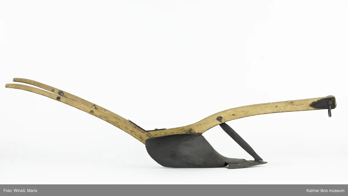 KLM 12823. Plog, trä, metall. Plog med handtag och ås av trä. På åsens ena sida sitter en rist av järn fastskruvad. Längst ut på åsen en dragkrok av järn för anspänning av dragdjur. Plogblad med bill av järn. Träkil mellan plogblad och landsida. Plogen är järnskodd på undersidan. Den har även varit järnskodd på landsidan men denna saknas. Spikarna sitter kvar i träet. Fyra slåar mellan handtagen, två av järn och två av trä. Det nedersta är fastskruvat i plogbladet. Plogen är svartmålad med en röd eventuell rostskyddsfärg under. Målad under senare tid.