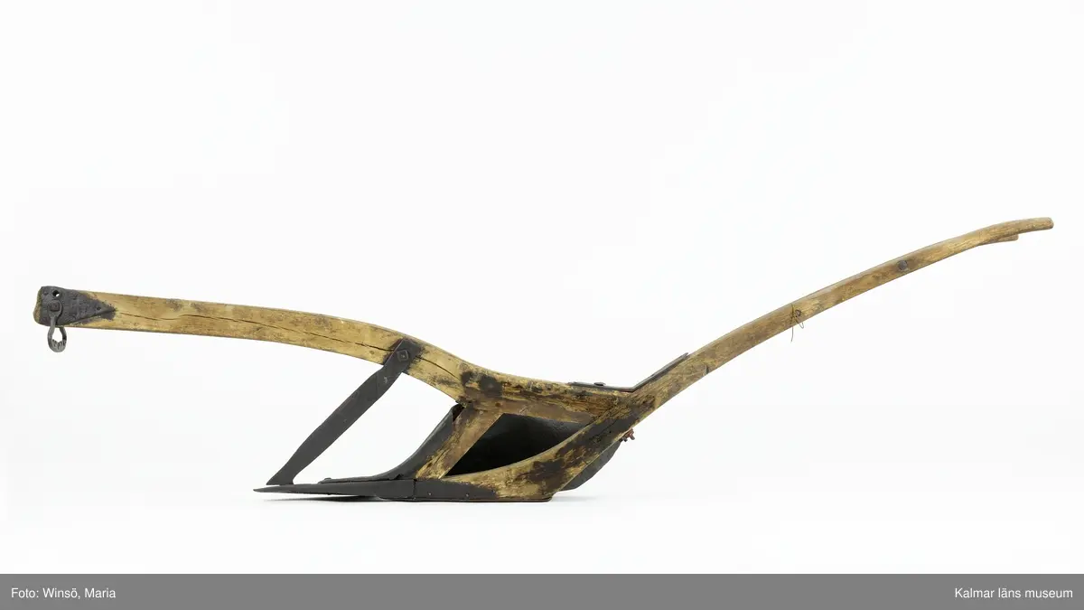 KLM 12823. Plog, trä, metall. Plog med handtag och ås av trä. På åsens ena sida sitter en rist av järn fastskruvad. Längst ut på åsen en dragkrok av järn för anspänning av dragdjur. Plogblad med bill av järn. Träkil mellan plogblad och landsida. Plogen är järnskodd på undersidan. Den har även varit järnskodd på landsidan men denna saknas. Spikarna sitter kvar i träet. Fyra slåar mellan handtagen, två av järn och två av trä. Det nedersta är fastskruvat i plogbladet. Plogen är svartmålad med en röd eventuell rostskyddsfärg under. Målad under senare tid.