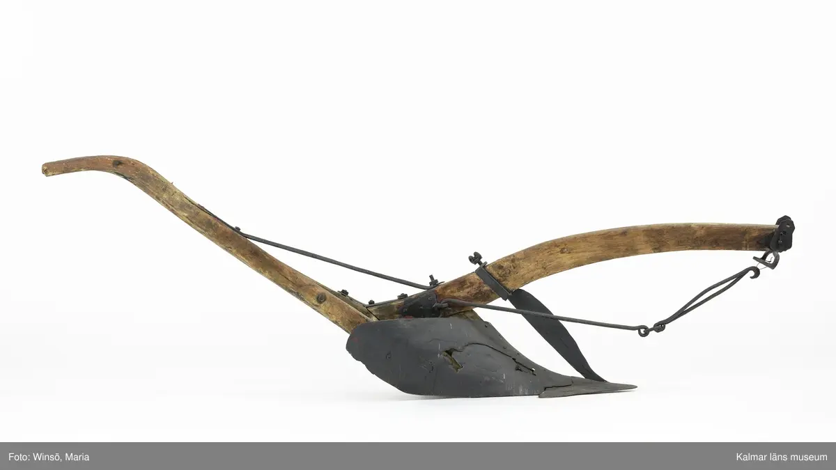 KLM 12824. Plog, trä, metall. Plog med handtag och ås av trä. Landsida av trä, järnskodd. Vändskiva och bill av järn. Tvärslå av järn mellan handtag och ås. Slåar mellan handtagen, två rundformade av järn, en U-formad av järn, en kantig av järn och en av trä. Längst fram på åsen en dragkrok av järn med reglage för inställning av höjd. På åsens mitt en fastskruvad rist. Risten är sammanfogad, eventuellt med färg, med billen. Tre långa krokar av järn hänger under åsen och möts i en järnring. Plogen är trasig på landsidan, träet är sprucket och järnbeslaget har lossnat. Plogen är rödmålad men färgen är kraftigt avskavd. Plogens järndetaljer är svartmålade med en röd eventuell rostskyddsfärg under. Den svarta färgen och rostskyddsfärgen är målad senare.