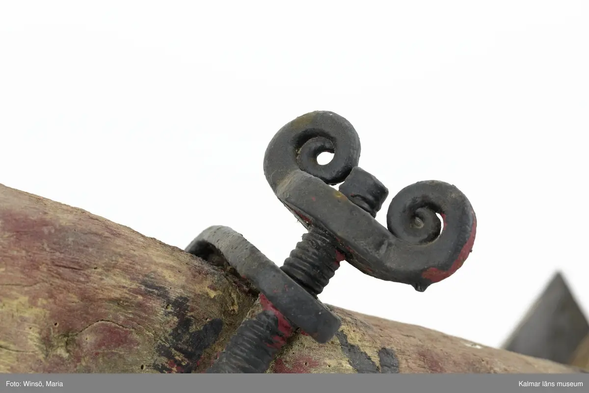 KLM 12824. Plog, trä, metall. Plog med handtag och ås av trä. Landsida av trä, järnskodd. Vändskiva och bill av järn. Tvärslå av järn mellan handtag och ås. Slåar mellan handtagen, två rundformade av järn, en U-formad av järn, en kantig av järn och en av trä. Längst fram på åsen en dragkrok av järn med reglage för inställning av höjd. På åsens mitt en fastskruvad rist. Risten är sammanfogad, eventuellt med färg, med billen. Tre långa krokar av järn hänger under åsen och möts i en järnring. Plogen är trasig på landsidan, träet är sprucket och järnbeslaget har lossnat. Plogen är rödmålad men färgen är kraftigt avskavd. Plogens järndetaljer är svartmålade med en röd eventuell rostskyddsfärg under. Den svarta färgen och rostskyddsfärgen är målad senare.