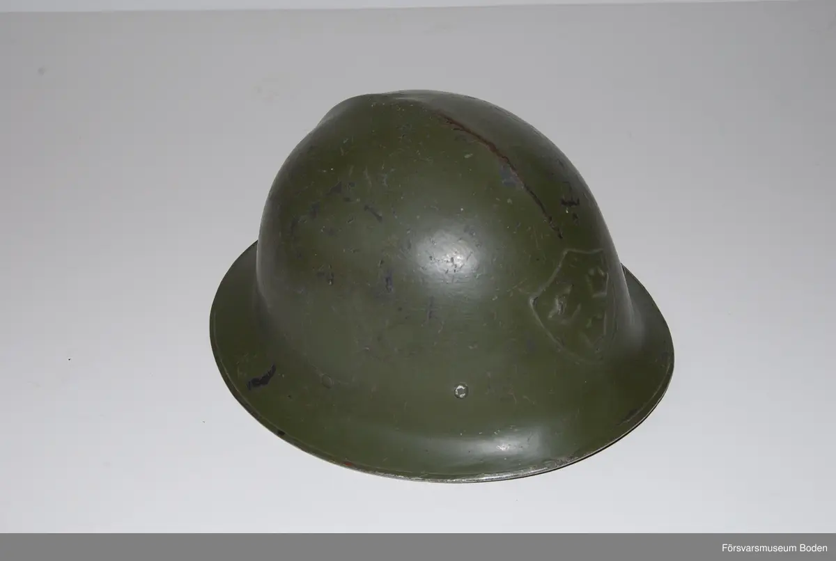 Olivgrön stålhjälm av typen med bredare och plattare brätte, den till vänster på bild 6 som jämför båda typerna av hjälm m/1921. Med insats bestående av tre läderkuddar som hålls ihop med skorem. Hakband av läder. Framtill tre kronor i relief, inga dekaler på sidorna. Upptill en förstärkningspressning i form av en kam.