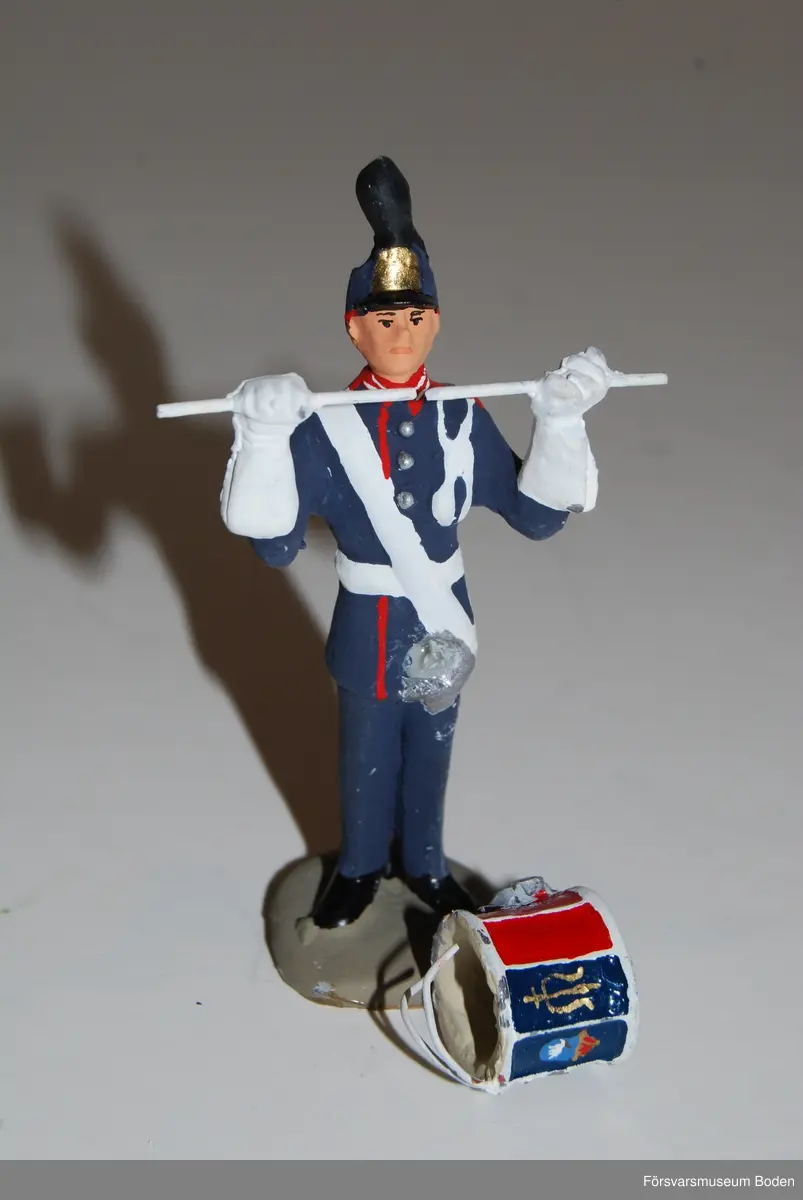 Gjuten och handmålad figur i skala 1:25. Förebilden är en trumslagare vid Arméns musikkår Norrland, Boden. Den pålimmade trumman har lossnat.