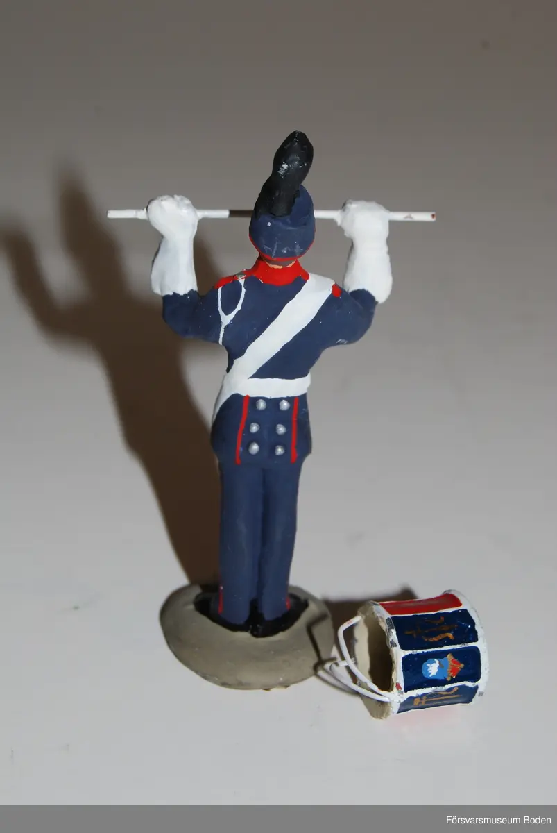 Gjuten och handmålad figur i skala 1:25. Förebilden är en trumslagare vid Arméns musikkår Norrland, Boden. Den pålimmade trumman har lossnat.