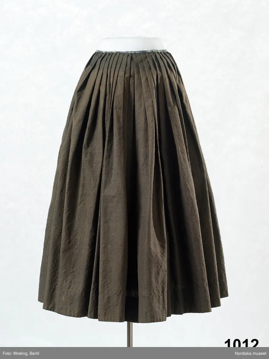 Kjol av gråsvart strävt och glansigt kamgarnstyg s.k. kamlott, veckad med motveck mitt bak, sprund vid vardera sidan om framvåden som har 3 veck åt vardera sidan, slät fram. Linning och 8 cm kantskoning av blå- och vitrutig linnelärft.

Använd som brudkjol midsommardagen 1795 av Annika Andersdotter. Hela bruddräkten inv.nr 1012 -1018. Även brudgummens dräkt bevarad inv.nr 1019- 1021. Se bilder i Anna-Maja Nyle´n: Folkdräkter, 1971.
Berit Eldvik okt 2005