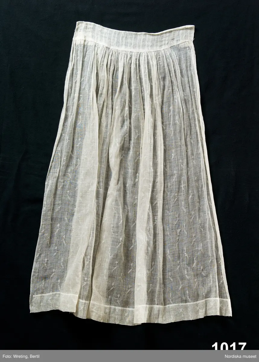 Förkläde av halvt genomskinligt tunt bomullstyg, s.k. linong med ett invävt mönster. Nedtill ca 5 cm bred fåll, upptill rynkat mot 6 cm bred och 40 cm lång linning av linong i annat mönster. Stadkant i sidorna.

Sådana tyger vävdes vid textilfabriker i Skottland i slutet av 1700-talet och kallas "lappet weaving" . Det gjordes med en speciell "lappet frame" en trälist lika bred som varpen och med en rad isatta nålar med öga i änden. Den satt under varpen och styrdes via en excentrisk skiva med mönstret inprogrammerat. Vissa av varptrådarna gick genom dessa ögon och när ramen fördes uppåt så att nålarna stack upp genom varpen och dessutom rördes i sidled genom skivans påverkan så bildade dessa varptrådar ett enkelt mönster på ytan . Mönstret ser ut att vara gjort med ett inplockat inslag, alltså i horisontell riktning men åstadkoms alltså med varptrådar som bringas att röra sig i "fel" riktning. 

Hör ihop med en hel bruddräkt inv.nr. 1012-1018, enligt traditionen använd på midsommardagen 1795 av bruden Annika Andersdotter, i Tunge socken, Ale hd, Västergötland.
Även brudgummens dräkt finns se inv.nr 1019 - 1021.
/Berit Eldvik aug 2005
Om förklädet verkligen bars redan 1795 är det ett mycket tidigt exempel på lappet weaving i bomull. 
/Berit Eldvik 2013-05-15