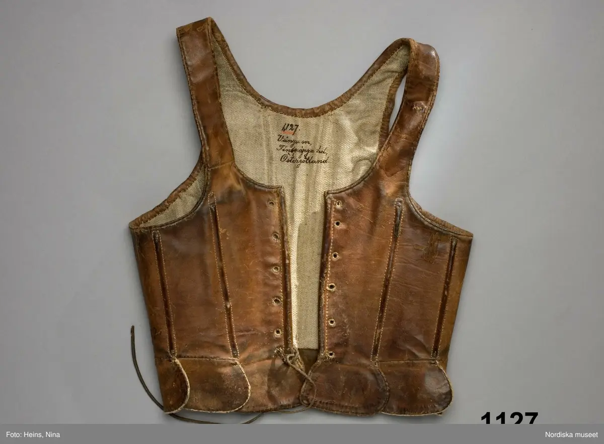 Katalogkort:  Lifstycke af brunt kalvskinn. Snörlif med insydda styfva spröt i rygg, sidor och framtill, sex par snörhål och en snörrem af skinn i det nedersta hålet på höger sida samt tio tegellagda flikar delvis i ett stycke med lifvet, delvis tillskarfvade. Foder af grov hvit linnehväf.
Tillhör den äldre kvinnodräkten, kalladt "munkalifstycke". Dylika lif, men af rödt kläde och med grönkantade flikar, förekommo i bruk långt in på 1840-talet.
/P.G.Vistrand/

Skinnet brunt, kraftigt. Med sammanlagt 12 insydda spröt  jämnt fördelade runt livet., snörhålen kantsydda med ljust lingarn. Foder av oblekt linne i spetskypert.
Anm. Helt men väl använt, nött och fläckigt.
Sådana skinnlivstycken användes framförallt till vardags, de var mycket slitstarka.
/Berit Eldvik 2008-02 15Kalvskinn.