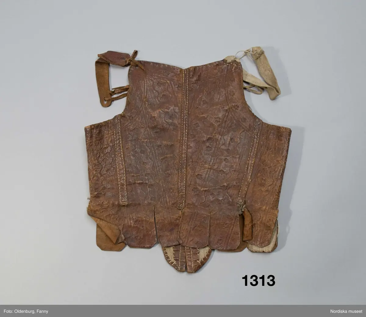 Katalogkort: "Lifstycke af brunt pressadt skinn. Öfver ryggsömmarna påsydda skinnremmar. Pressningen utgöres af ränder, våglinier, små rosetter m.m. Foder af hvit, grof linneväf. Å insidan af ena bröststycket märkt: "Segrin" med bläck. Till kvinnodräkt, enl. uppgift från 1700-talet."/P.G.Vistrand