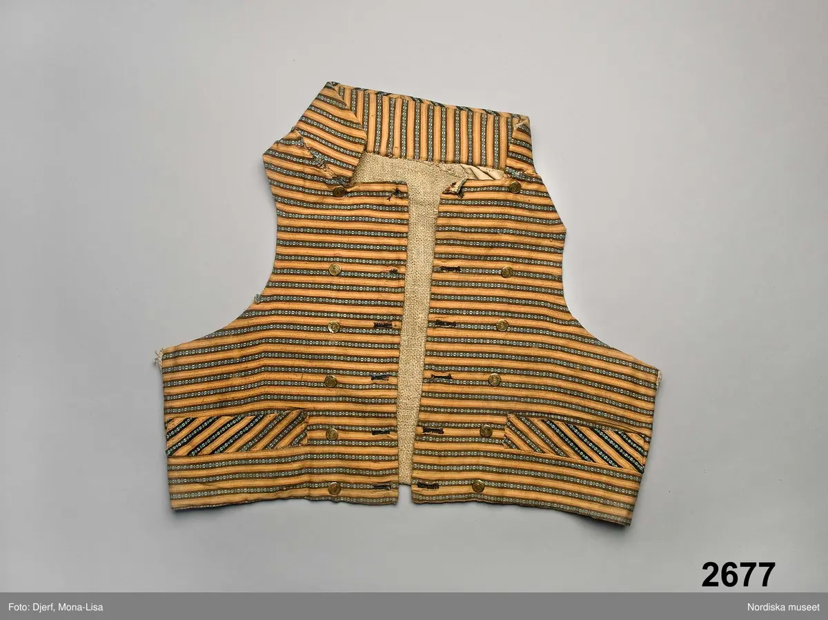 Väst i kort modell av randig bomullsrips i brunt och beige med invävd mönsterrand i den mörkbruna randen i ljusblått med vita "hoppor". Västen är tvärrandig med 2 framstycken och hög styvad  krage av samma tyg, ryggstycke av fin oblekt linnelärft, foder av grov och gles linnelärft i ryggen och finare i framstyckena, 2 stolpfickor, dubbelknäppning med knapphål på båda framstyckena och 6 par mässingsknappar med Ångermanlands landskapsvapen med 3 liggande laxar.
Tygsorten finns under benämningen: Västtyg i flera av 1820-30 talets tygprovsböcker och har enligt uppgift använts som brudgumsväst 1824
/Berit Eldvik  2005-11