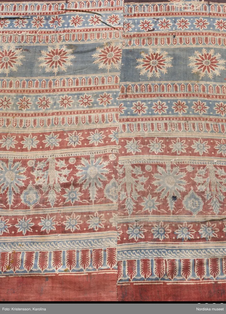 Stort tygstycke sammansytt av 3 våder bomullslärft  á 54 cm bredd. Hopsydda med vitt handspunnet lingarn. Tryckt mönster i rött och blått på vitt underlag.. I mitten stort cirkelmotiv fylld med mönster och i mitten en stjärna, åt fyra håll en triangel ut från cirkeln och mot 4 större snäckliknande ornament. I varje hörn motiv med 4 fiskar med stjärtarna in mot en cirkel, Kantbårder bestånde av olikmönstrade ränder i olika bredder.  Trycket är gjort med tryckstockar och smalare tryckstämplar i kantbårderna där tydliga blockskarvar syns.  Tekniken är den gamla indiska med betmålning av de partier som skall bli röda och därefter doppning i krappbad, tvättning av tyget före blåfärgning som görs så att vita och röda parteier täcks med en pasta före färgning i indigokyp. På baksidan är tyget mycket blåare än på framsidan. Spegelns botten har färgats först röd och därefter blå  så att den fått en mörklila , lätt flammig nyans. Tyget är skört och trasigt på flera ställen, längs hela högersidan är det söndigt vilket kan vara musätet. En del mindre hål har lappats med  gråblått siden på baksidan eller på ett ställe med en bit av själva tygtrycket. Även blekt.
Insamlat av Nils Månsson-Mandelgren vid hans resor i Västergötland, inga uppgifter finns.
 /Berit Eldvik 2013-06