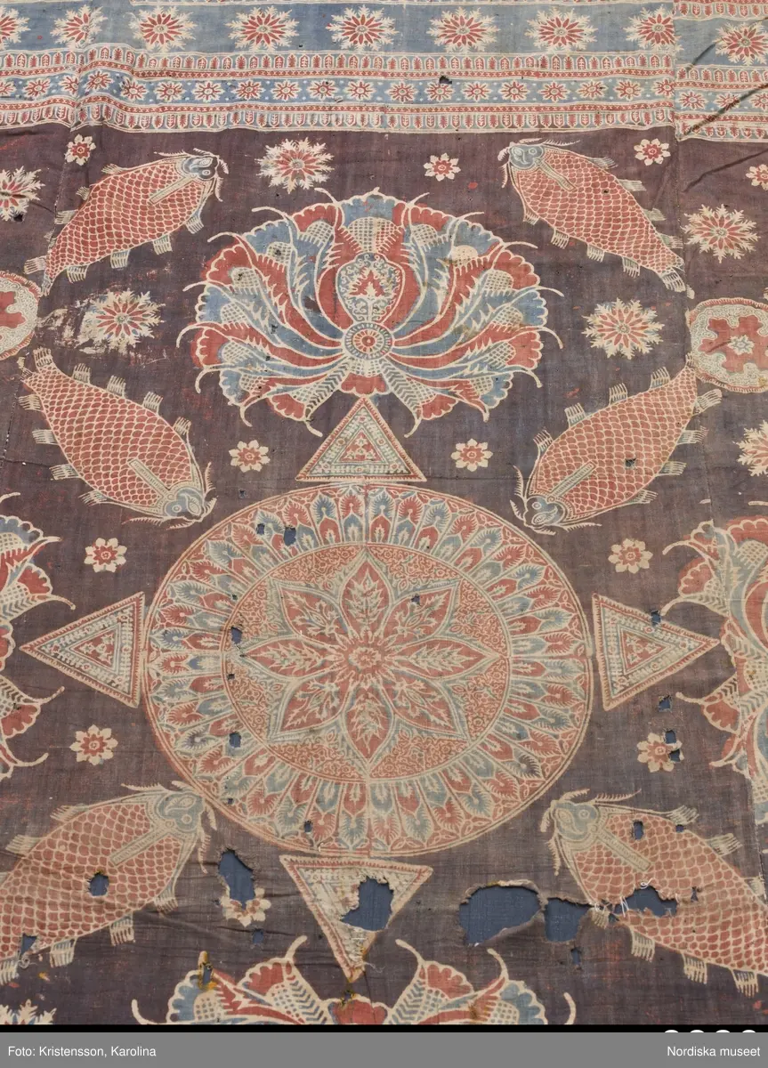 Stort tygstycke sammansytt av 3 våder bomullslärft  á 54 cm bredd. Hopsydda med vitt handspunnet lingarn. Tryckt mönster i rött och blått på vitt underlag.. I mitten stort cirkelmotiv fylld med mönster och i mitten en stjärna, åt fyra håll en triangel ut från cirkeln och mot 4 större snäckliknande ornament. I varje hörn motiv med 4 fiskar med stjärtarna in mot en cirkel, Kantbårder bestånde av olikmönstrade ränder i olika bredder.  Trycket är gjort med tryckstockar och smalare tryckstämplar i kantbårderna där tydliga blockskarvar syns.  Tekniken är den gamla indiska med betmålning av de partier som skall bli röda och därefter doppning i krappbad, tvättning av tyget före blåfärgning som görs så att vita och röda parteier täcks med en pasta före färgning i indigokyp. På baksidan är tyget mycket blåare än på framsidan. Spegelns botten har färgats först röd och därefter blå  så att den fått en mörklila , lätt flammig nyans. Tyget är skört och trasigt på flera ställen, längs hela högersidan är det söndigt vilket kan vara musätet. En del mindre hål har lappats med  gråblått siden på baksidan eller på ett ställe med en bit av själva tygtrycket. Även blekt.
Insamlat av Nils Månsson-Mandelgren vid hans resor i Västergötland, inga uppgifter finns.
 /Berit Eldvik 2013-06