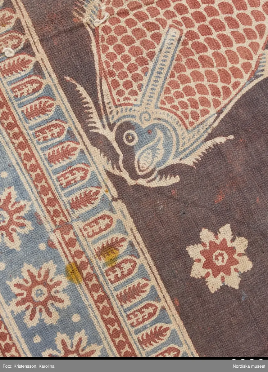 Stort tygstycke sammansytt av 3 våder bomullslärft  á 54 cm bredd. Hopsydda med vitt handspunnet lingarn. Tryckt mönster i rött och blått på vitt underlag.. I mitten stort cirkelmotiv fylld med mönster och i mitten en stjärna, åt fyra håll en triangel ut från cirkeln och mot 4 större snäckliknande ornament. I varje hörn motiv med 4 fiskar med stjärtarna in mot en cirkel, Kantbårder bestånde av olikmönstrade ränder i olika bredder.  Trycket är gjort med tryckstockar och smalare tryckstämplar i kantbårderna där tydliga blockskarvar syns.  Tekniken är den gamla indiska med betmålning av de partier som skall bli röda och därefter doppning i krappbad, tvättning av tyget före blåfärgning som görs så att vita och röda parteier täcks med en pasta före färgning i indigokyp. På baksidan är tyget mycket blåare än på framsidan. Spegelns botten har färgats först röd och därefter blå  så att den fått en mörklila , lätt flammig nyans. Tyget är skört och trasigt på flera ställen, längs hela högersidan är det söndigt vilket kan vara musätet. En del mindre hål har lappats med  gråblått siden på baksidan eller på ett ställe med en bit av själva tygtrycket. Även blekt.
Insamlat av Nils Månsson-Mandelgren vid hans resor i Västergötland, inga uppgifter finns.
 /Berit Eldvik 2013-06