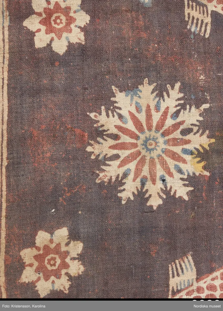 Stort tygstycke sammansytt av 3 våder bomullslärft  á 54 cm bredd. Hopsydda med vitt handspunnet lingarn. Tryckt mönster i rött och blått på vitt underlag.. I mitten stort cirkelmotiv fylld med mönster och i mitten en stjärna, åt fyra håll en triangel ut från cirkeln och mot 4 större snäckliknande ornament. I varje hörn motiv med 4 fiskar med stjärtarna in mot en cirkel, Kantbårder bestånde av olikmönstrade ränder i olika bredder.  Trycket är gjort med tryckstockar och smalare tryckstämplar i kantbårderna där tydliga blockskarvar syns.  Tekniken är den gamla indiska med betmålning av de partier som skall bli röda och därefter doppning i krappbad, tvättning av tyget före blåfärgning som görs så att vita och röda parteier täcks med en pasta före färgning i indigokyp. På baksidan är tyget mycket blåare än på framsidan. Spegelns botten har färgats först röd och därefter blå  så att den fått en mörklila , lätt flammig nyans. Tyget är skört och trasigt på flera ställen, längs hela högersidan är det söndigt vilket kan vara musätet. En del mindre hål har lappats med  gråblått siden på baksidan eller på ett ställe med en bit av själva tygtrycket. Även blekt.
Insamlat av Nils Månsson-Mandelgren vid hans resor i Västergötland, inga uppgifter finns.
 /Berit Eldvik 2013-06