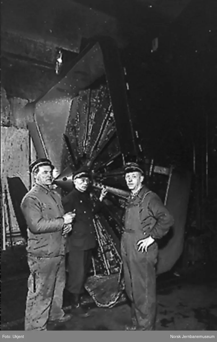 Tre mann foran en roterende snøplog, trolig inne i en lokomotivstall