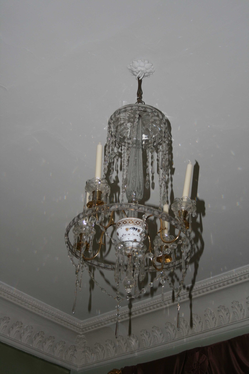 1 prismelysekrone, Louis seize, ca 1790.
Hjulkrone.
4 messingarmer m/skåler og prismeanheng, tøndebånd.  Glasslokk øverst, glass-stang i midten og hvit porselen m /gulldekor, nederst behengt med prismer.
Bømisk krone (iflg. eldre protokoll)
Lysarmene i messing er sekundære, det skulle vært glassarmer.