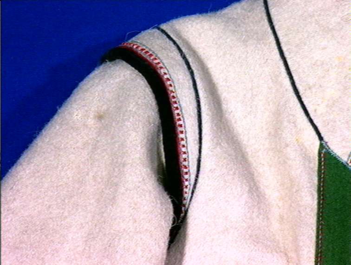 Hvit mannskofte av vadmal med gønne kanter av klede (oppslag).
Nedrekanter, ermebryninger og halskrage har dekor av rød korstign avgrenset av blått konturstign (ullgarn).
Halskrage ha kanting av svartklede med tittkant av rødt klede.
Ermer har et ca. 6,5 cm bredt belegg av grønn klede kantet med rødt klede.