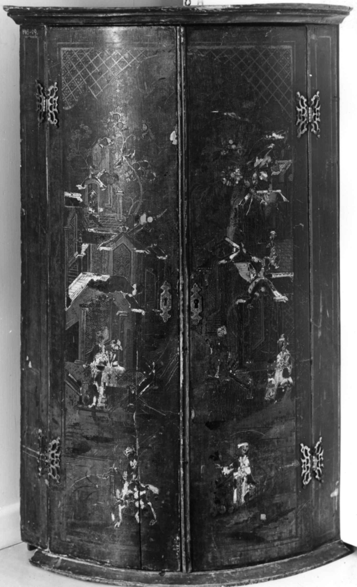 Hjørneskap med buet frontside. To dører med asiatisk lakkteknikk, fremstilling av hus, planter, figurer i flatrelieff. Innvendig 3 hyller.