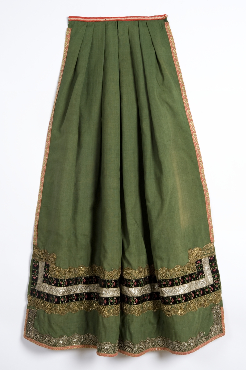 Grønt forkle av ull med dekor av metallblonder og bånd.