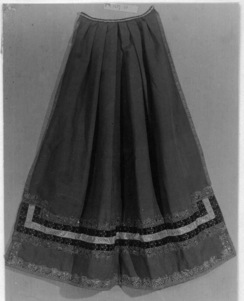 Grønt forkle av ull med dekor av metallblonder og bånd.