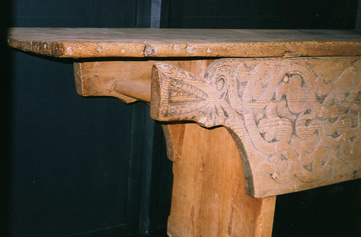 Langbord med forvaringsrom bak høye sarger. Fremsarg er med heldekkende flatskurd ranker og drageornamentikk samt en luke til forvaringsrommet. 