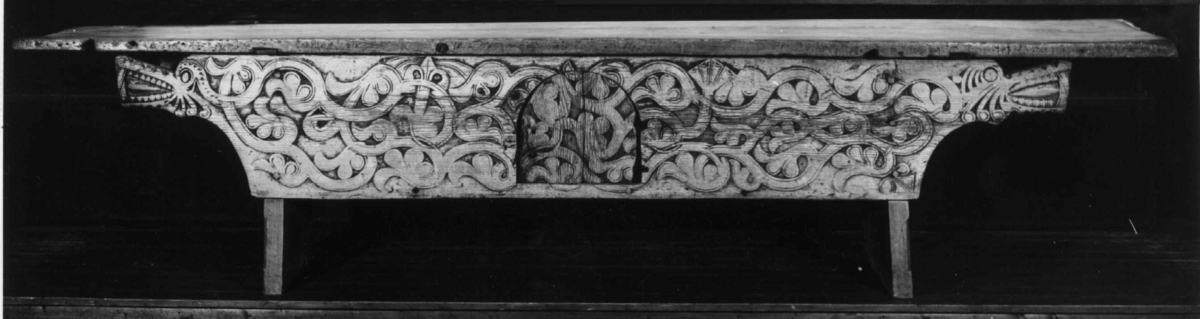 Langbord med forvaringsrom bak høye sarger. Fremsarg er med heldekkende flatskurd ranker og drageornamentikk samt en luke til forvaringsrommet. 