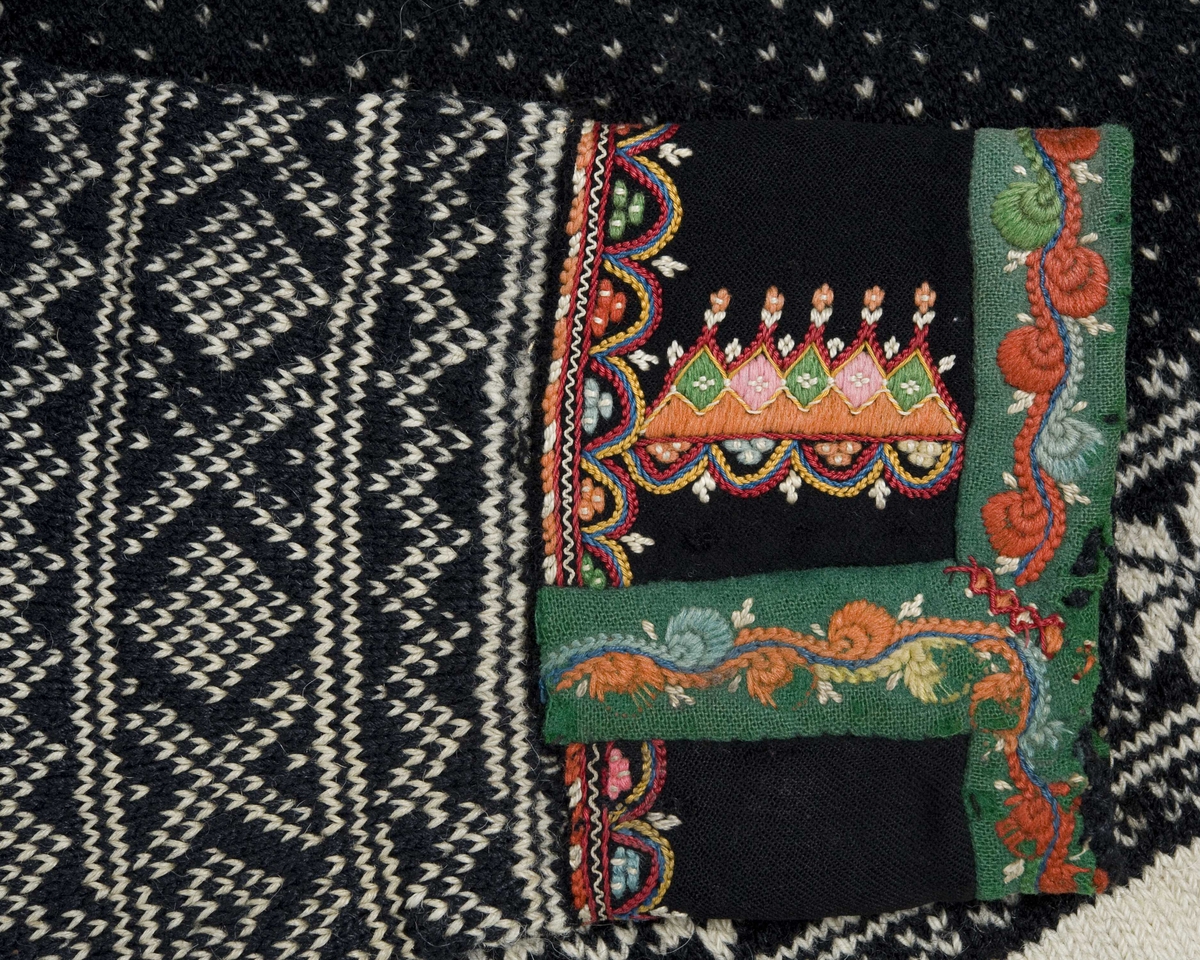 Strikket ullgarn, heimespunnet, svart og hvitt. Broderi i ullgarn. Langs nedrekanten en 13 cm bred hvit bord, resten mønsterstrikket: en bord åttebladroser, en sikksakkbord, en bord kors og kantstilte kvadrat. Langs åpningen foran og halsringingen er påsatt et stykke svart klede, saum i rosa, blått, gult og grønt zephyrgarn. Rundt ermet et 9,3 cm bredt belegg av svart klede, kantet med grønn klede 2,5 cm bred. En brodert bord langs indre og ytre kant av belegget. Aagot Noss 1969. 
