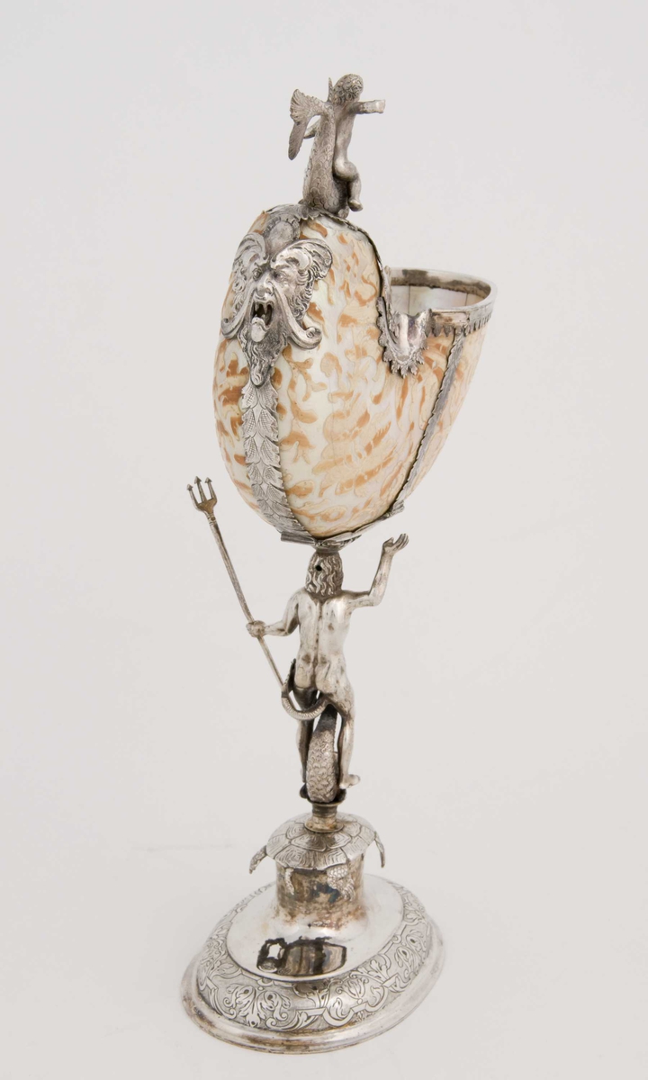 Skall av blekksprutarten Nautilus. Skåret dekorasjon, fot og innfatning sølv, støpt og gravert dekorasjon. Stpl: MF/H//1720 Morten Finchenhagen, Christiania. Skallet er brunflammet i overflaten. Nautilusblekkspruten lever i sydlige hav omkring Filippinene.