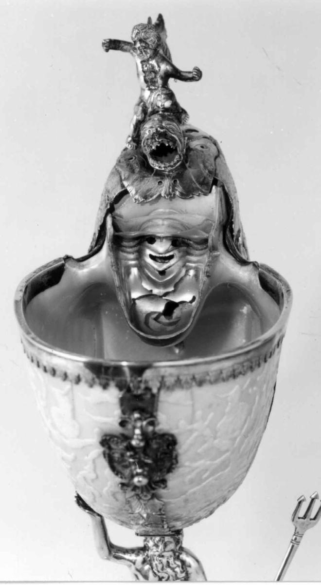 Skall av blekksprutarten Nautilus. Skåret dekorasjon, fot og innfatning sølv, støpt og gravert dekorasjon. Stpl: MF/H//1720 Morten Finchenhagen, Christiania. Skallet er brunflammet i overflaten. Nautilusblekkspruten lever i sydlige hav omkring Filippinene.