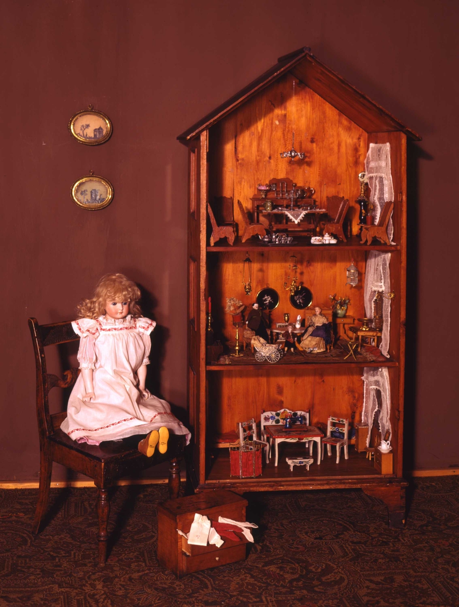 Dukkehus i tre. Huset har 3 etasjer og et spisstak. Hver etasje har 2 vinduer med gardiner. Overflaten er beiset og lakkert.