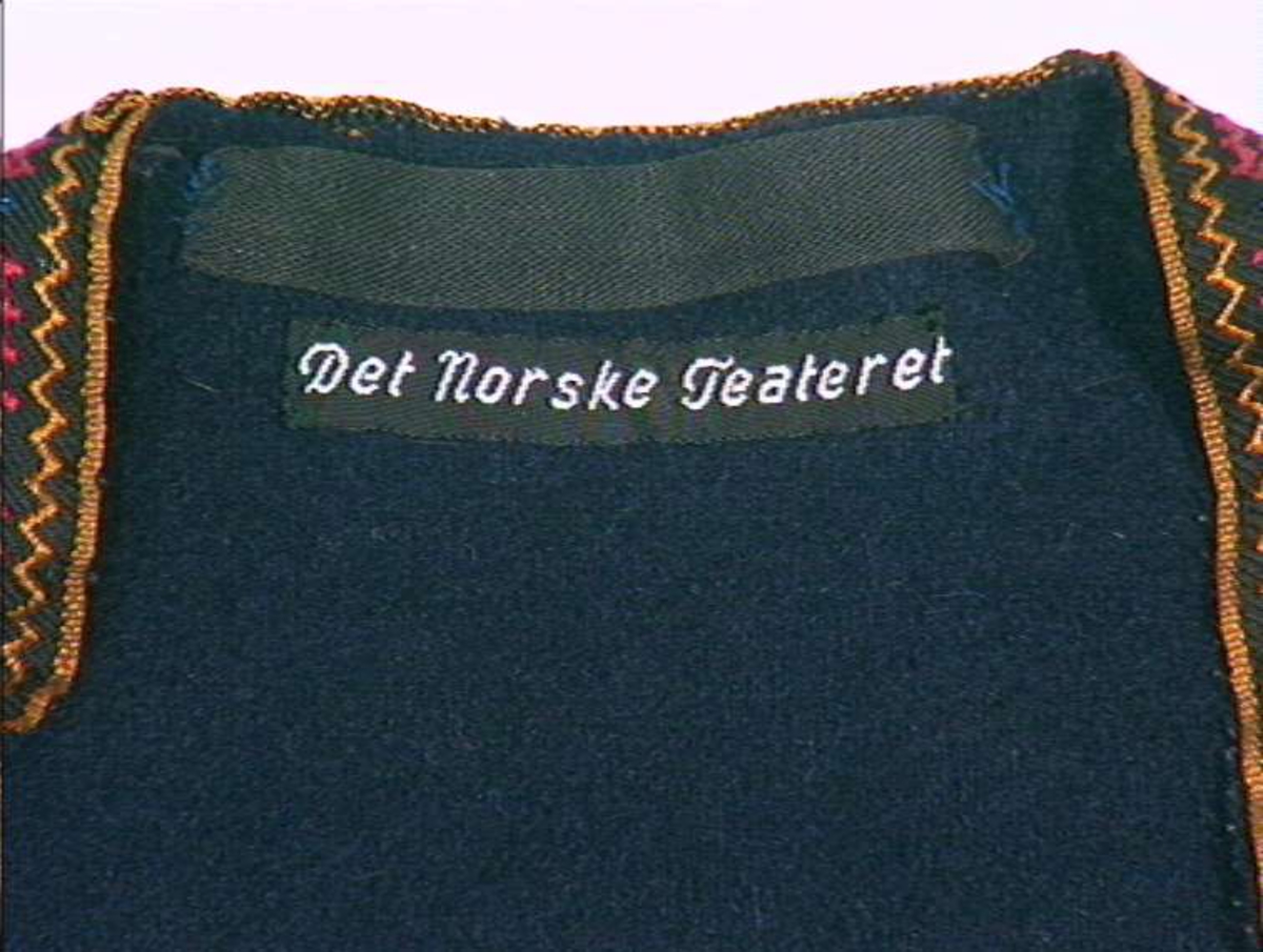 Mørkeblå trøye i ull, maskinsydd. Dekor av 3 cm brede bånd langs fremkanter, hals og ermeringning.I rygg nederst er det 3 innfeldte trekanta kiler som er foret med linlerret og bomullsstoff.