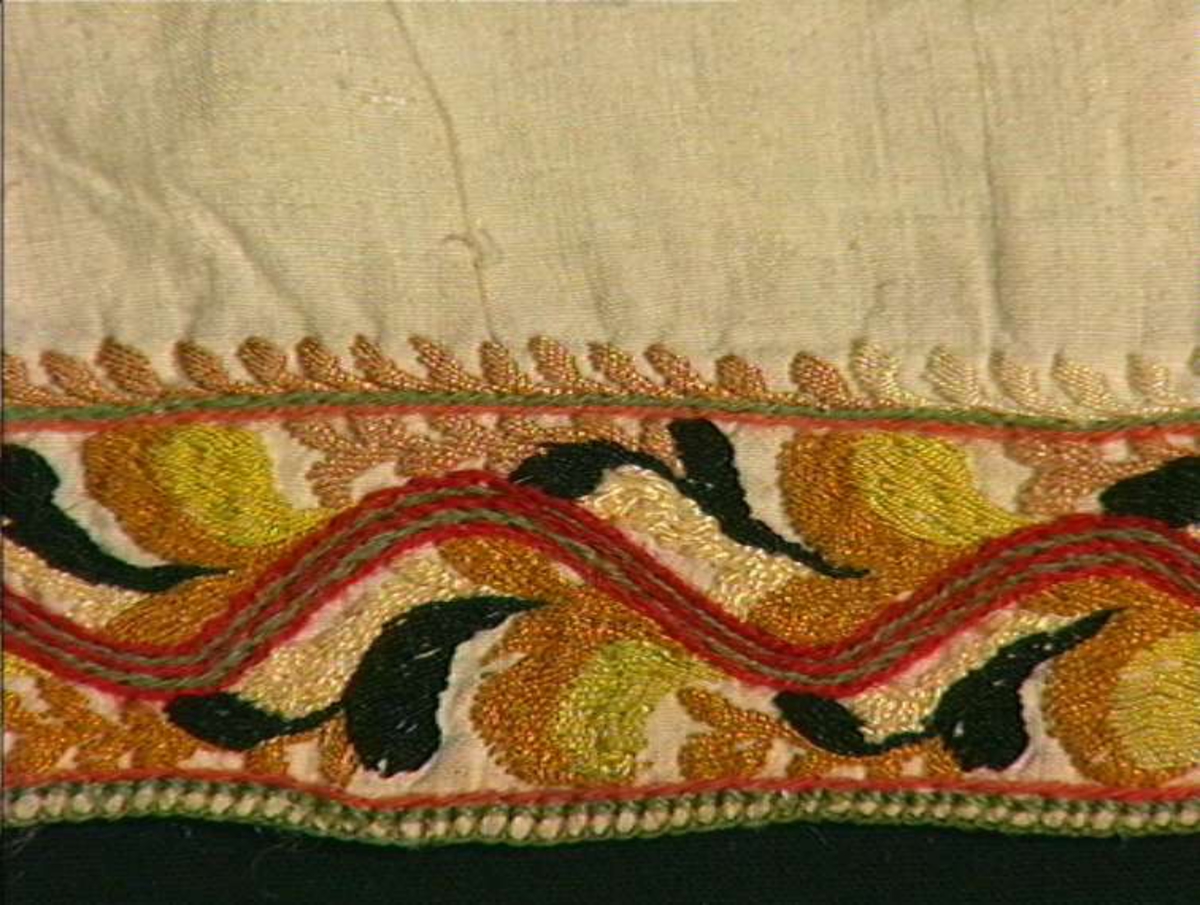 Skjorte fra Sauland, Hjartdal. Broderi på bolen uttrekksøm, klostersøm og korssting. Halslinning og slengerm sydd i plattsøm 
Halskvarde (halslinning) mål:  7,7cm.x57cm ca. Broderi 6,8cm.
Broderi på skulder ca 5,5x6,5cm. 
Brodert bord på slengerm 4cm., avslutning knapphullskant og attersting.