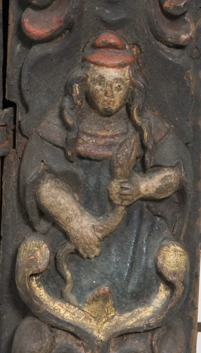 Skap med to dører. Skåret dekor, tre firgurer, en med slange i hendene, en med hendene i bedeposisjon, en med anker – representerer trolig hhv. visdom, tro og håp og visdom (tre av de syv dyder).