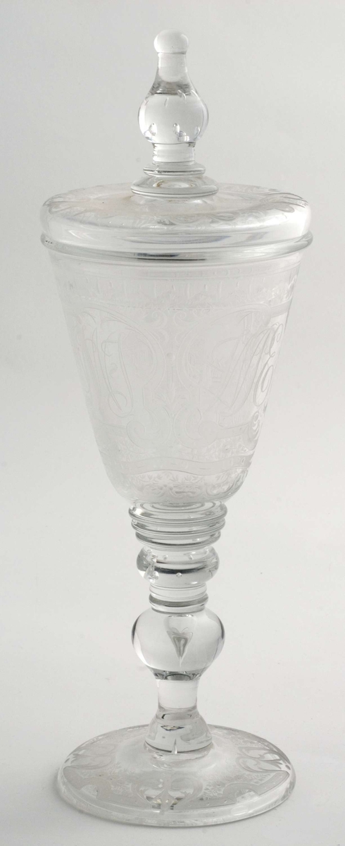 Pokal med lokk, glass. Fot har graverte blomster  og knekkede bånd. På cupa gravert bilde av Bogstad på en side, på den andre: N.W.J.  W.P.E.