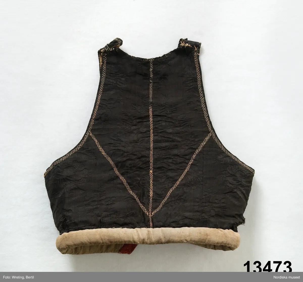 Livstycke av svart sidendamast, 2 framstycken, 1 bakstycke med flätsömmar i gulrosa silke som markerar "sömmar" i ryggen och som omger ärmhålen. På framstyckena broderier i stjälksöm, och flätsöm. Framkanterna infodrade med remsa av röd vadmal. Snörning med 5 par maljor som på ena stycket har hjärtformigt fäste fastsydda med svart lintråd och på det andra cirkelformigt sydda med rött ullgarn, nederst hyska och hake, foder och valk av oblekt linnelärft.
Anm. Mycket slitet och sidenet delvis sprucket.

/Berit Eldvik maj 2006
