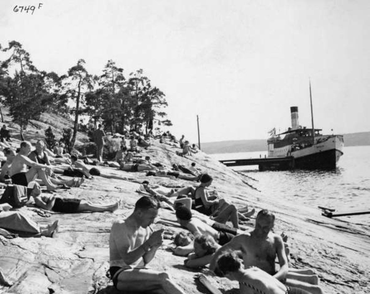 Strandliv med badegjester som soler seg på svaberget ved sjøen på Ingierstrand bad utenfor Oslo. Båt ved brygga i bakgrunnen.