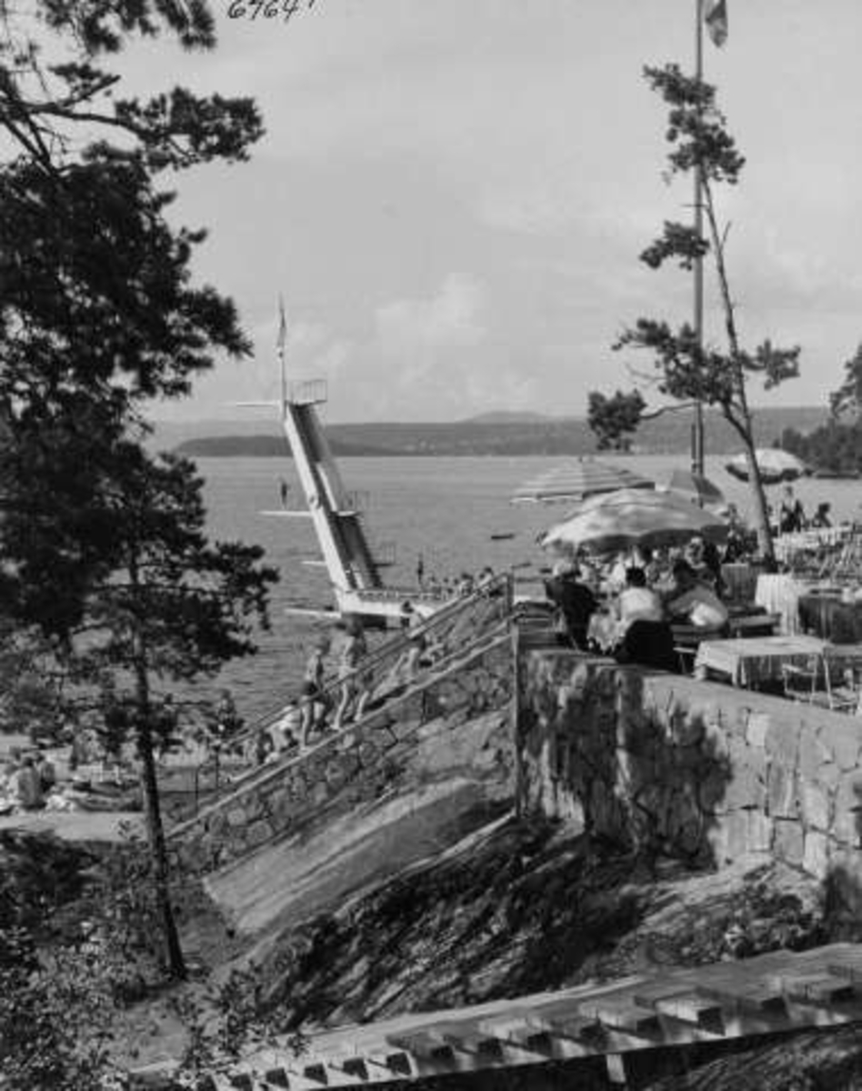 Strandliv med badegjester  ved sjøen på Ingierstrand bad utenfor Oslo. Stupetårn i bakgrunnen for terrassen med kafé og gjester under parasoller.