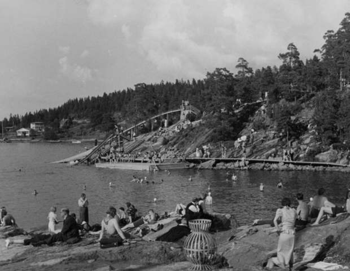 Strandliv med badegjester som soler seg på svaberget ved sjøen på Ingierstrand bad utenfor Oslo. Vannsklie i bakgrunnen