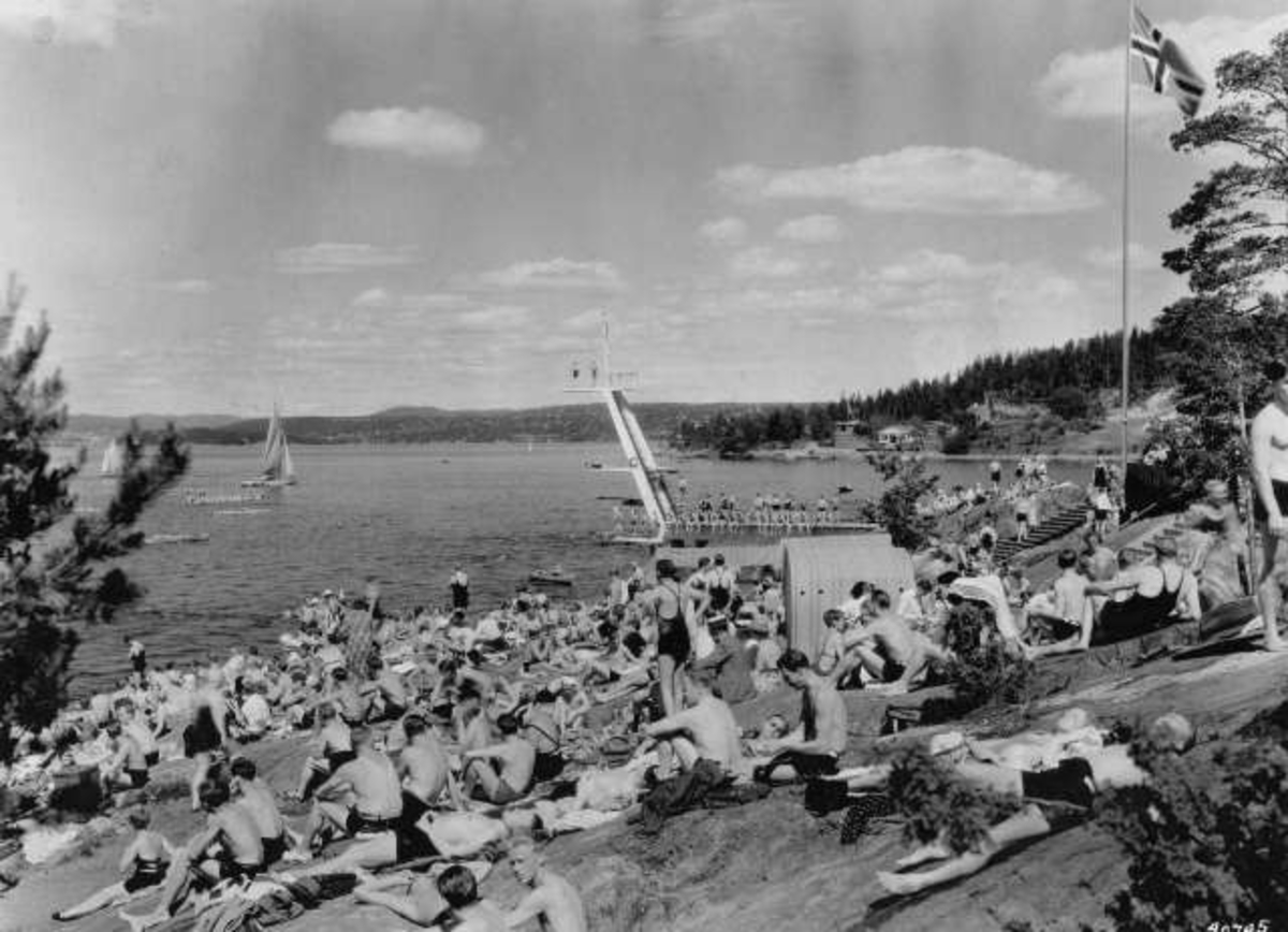 Strandliv med badegjester som soler seg på svaberget ved sjøen på Ingierstrand bad utenfor Oslo. Stupetårn i bakgrunnen.