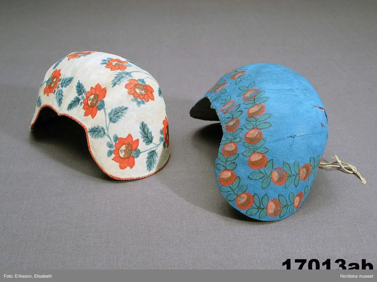 Huvudliggaren bilaga= "Två hvardagsmössor" från Härja socken i Vestergötland, såsom de allmänt begagnades på 1830-talet af gifta qvinnor."

a. Bindmössa, mått från öra till öra 35 cm, djup 11 cm.
b. Bindmössa längd från öra till öra 37 cm

a. av vitt stärkt bomullstyg i panama sydd över en stomme av grå lumppapp, foder saknas. Blommönster i rött och blåttoch något gult  är schablonmålat på tyget efter det att tyget är monterat.Tunn målad röd kant. Tunt dragsnöre i nacken.

b. lik a. av tyg målad i blått med 2 rader blommotiv i rött och grönt och något svart, inuti märkt M med bläck.Knytband i nacken.


Berit Eldvik mars-2005